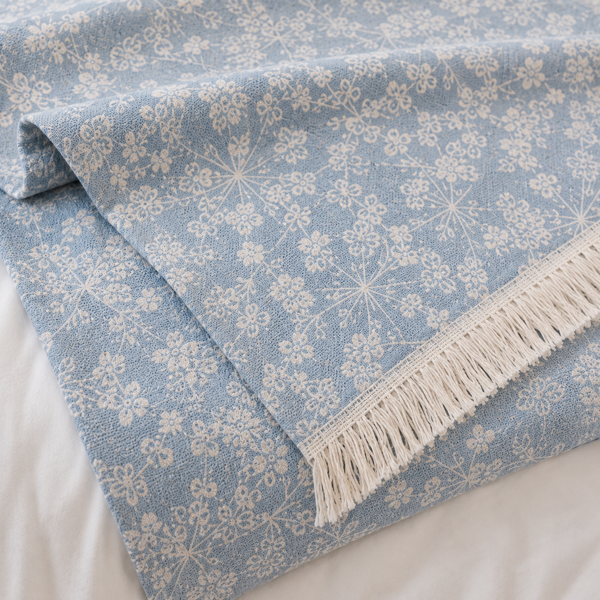 A folded light blue woven textile with an off-white floral snowflake pattern and cream fringe, shown on a white fabric background.