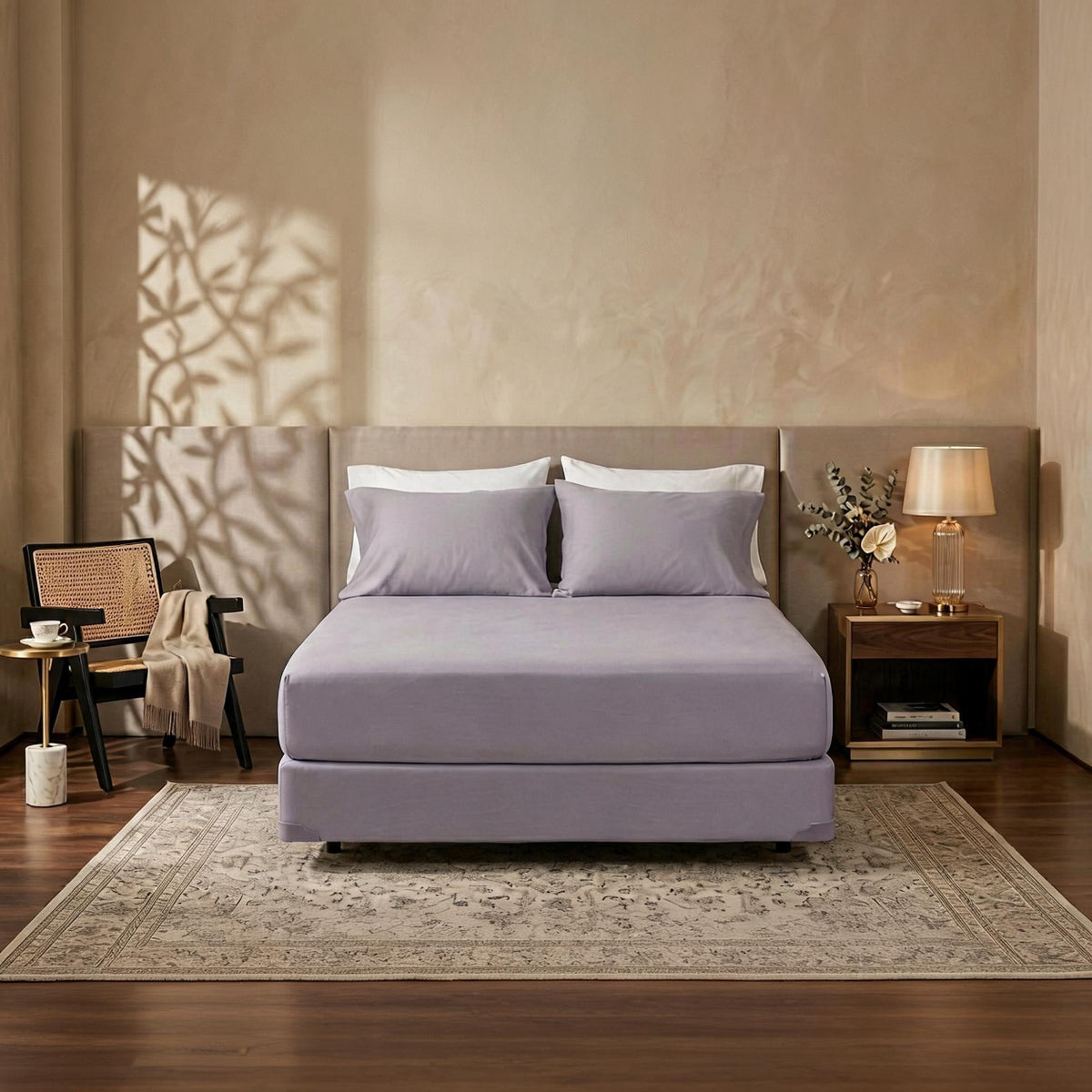 A bed dressed in a light lavender bedding set with two white pillows, styled in a warm, neutral-toned bedroom with a wooden floor, patterned rug, and bedside table.