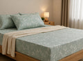 A bed dressed in a light sage green sheet set with a delicate white floral pattern, styled with matching pillowcases and a cream-colored ribbed throw blanket in a softly lit bedroom.