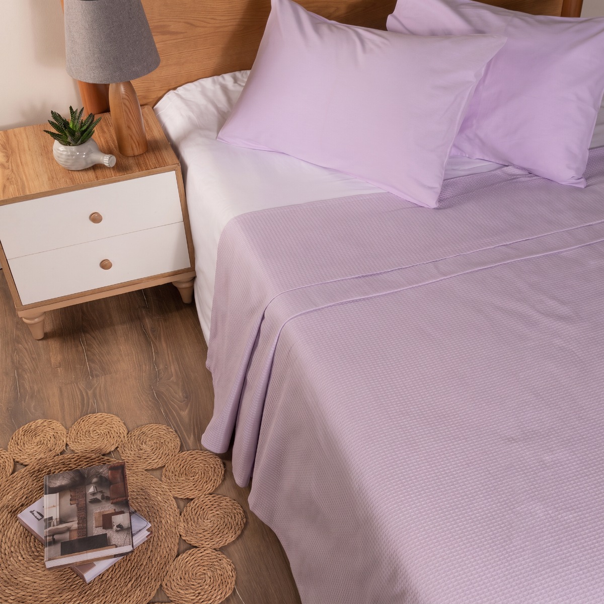 A neatly made bed with light lavender bedding, including a textured coverlet and matching pillowcases, styled in a bedroom setting with a wooden nightstand, lamp, and woven floor accents.